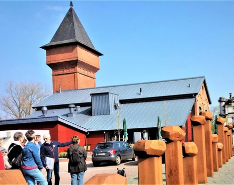 Eisenhütte-Puddelöfen-Augustfehn-Apen Eine Gruppe von Menschen steht vor einem historischen Backsteinbau mit einem großen Turm und modernen metallischen Akzenten. Ein blauer Himmel und zahlreiche Metallskulpturen sind im Vordergrund sichtbar.