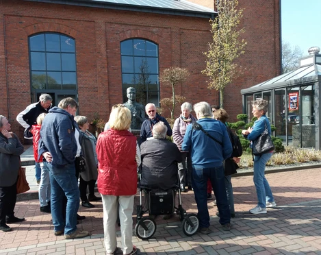 Gästeführung an der Eisenhütte Eine Gruppe von Personen nimmt an einer Führung vor einer historischen Backsteinfassade teil, darunter eine Metallbüste.