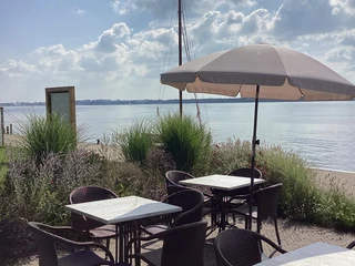 Terrasse an Anleger Insel Wilhelmstein Terrassenmöbel mit Blick auf den See und Segelmast, sanft beleuchtet von der Sonne, unter blauem Himmel.