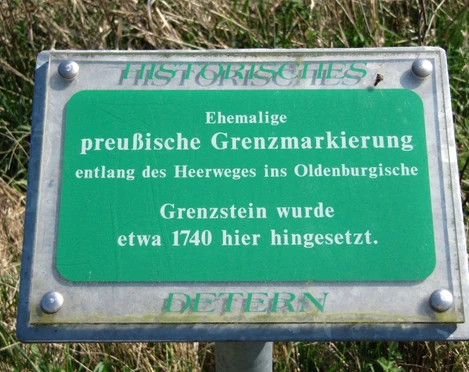 Grenzstein Holtgast Ein grünes Schild mit der Aufschrift "Ehemalige preußische Grenzmarkierung" steht in einer Wiese. Der Text erklärt, dass der Grenzstein entlang des Heerweges im Oldenburgischen etwa 1740 hier aufgestellt wurde. Das Schild ist mit "Historisches Deterner" gekennzeichnet.