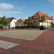 rathausmarkt-apen Der Rathausmarkt Apen zeigt eine harmonische städtische Szenerie mit roten Pflastersteinen und Gebäuden im traditionellen Baustil. Ein blauer Himmel und grüne Bäume ergänzen das Bild.
