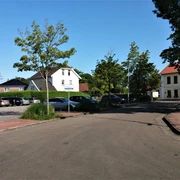 Viehmarktplatz-in-Apen Der Viehmarktplatz in Apen zeigt eine ruhige Straßenszene. Links parken Autos vor grünen Hecken und einem roten Dach. Im Hintergrund gibt es Bäume und der Himmel ist blau. Rechts steht ein hellgelbes Gebäude mit einem roten Dach.