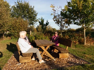 Gäste auf dem Rastplatz Hengstforde Zwei ältere Personen und eine jüngere Frau sitzen entspannt an einem Holztisch auf dem Rastplatz Hengstforde, umgeben von Bäumen und Wiesen im warmen Sonnenlicht.