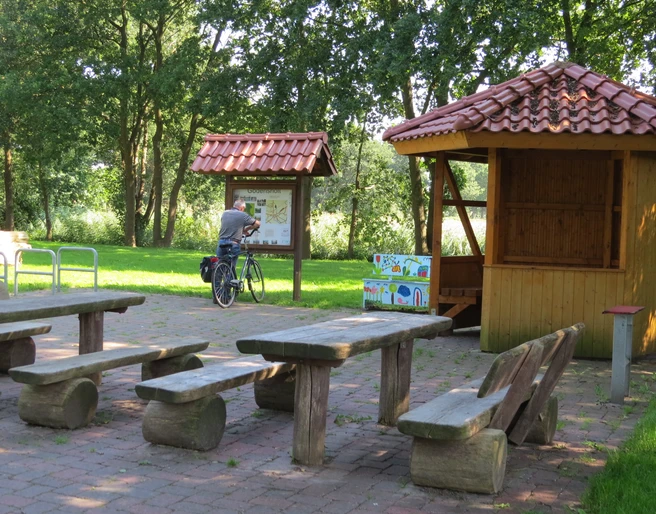 Rastplatz Am Drakamp Holzpicknicktische stehen auf gepflastertem Gelände, flankiert von einer Info-Tafel und einem Radfahrer.