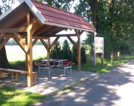 Rastplatz Alter Kirchweg Ein hölzerner Unterstand mit rotem Ziegeldach bietet Schutz, umgeben von Bäumen und neben einem Weg.