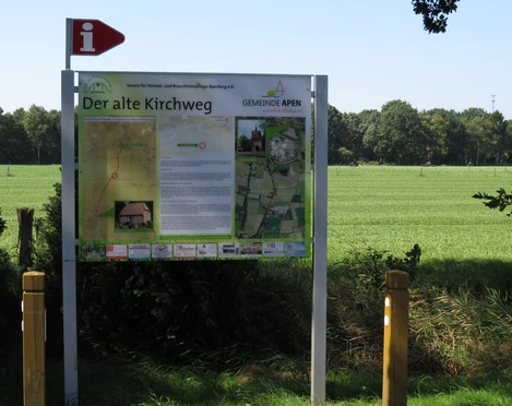 Infotafel Alter Kirchweg Die Infotafel "Alter Kirchweg" steht vor einer grünen Wiese und beschreibt die Umgebung und Historie des Wanderweges. Im Hintergrund ist eine Baumreihe sichtbar, die die natürliche Landschaft prägt.