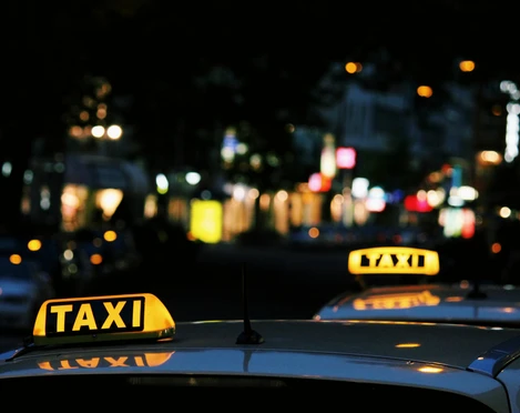 taxi Im Bild sind zwei Taxis mit leuchtendem Taxi-Schild auf dem Dach in einer belebten nächtlichen Straße zu sehen.