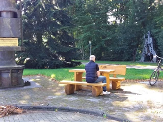 Rastplatz am Stahlwerk Augustfehn II Ein älterer Mann sitzt auf einer Holzbank an einem Rastplatz umgeben von hohen Bäumen. Ein Fahrrad lehnt an der Bank.
