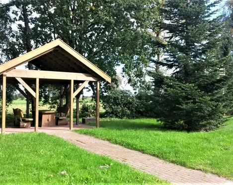Rastplatz Bokelermoor Ein überdachter Rastplatz auf grüner Wiese, umgeben von Bäumen, mit einem gepflasterten Weg.