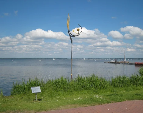Eine Skulptur mit Kurven und Kugeln steht am Seeufer, segelnde Boote und Wolken im Hintergrund.