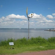 Gesamtansicht Skulptur Tanz der Winde Eine Skulptur mit Kurven und Kugeln steht am Seeufer, segelnde Boote und Wolken im Hintergrund.