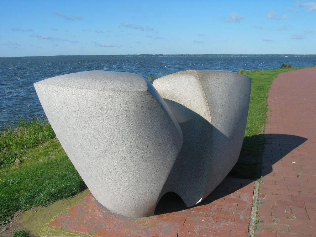 Swingende Welle Moderne Skulptur aus Stein, bestehend aus geschwungenen Formen, steht vor einem See mit blauem Himmel.