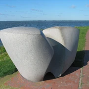 Swingende Welle Moderne Skulptur aus Stein, bestehend aus geschwungenen Formen, steht vor einem See mit blauem Himmel.