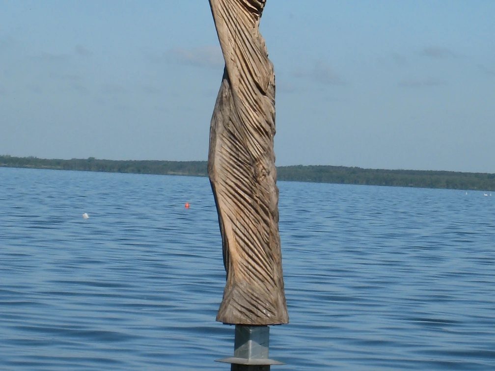 Skulptur aus Holz im Wasser, mit kunstvollen Falten, die dem Wind nachempfunden sind, vor blauem Himmel.