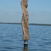 Skulptur aus Holz im Wasser, mit kunstvollen Falten, die dem Wind nachempfunden sind, vor blauem Himmel.