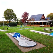Minigolf Goldener Hecht Ein weitläufiger Minigolfplatz mit verschiedenen Bahnen, Bänken und einem hölzernen Pavillon im Hintergrund.
