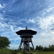 Ein hölzerner Aussichtsturm erhebt sich unter einem blauen Himmel mit verstreuten Wolkenformationen.