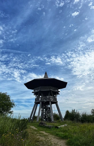 Ein hölzerner Aussichtsturm erhebt sich unter einem blauen Himmel mit verstreuten Wolkenformationen.