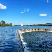 Segelboot gleitet auf dem sonnenbeschienenen Lippesee nahe eines runden, künstlichen Wasserfalls.