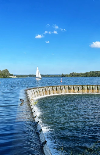 Segelboot gleitet auf dem sonnenbeschienenen Lippesee nahe eines runden, künstlichen Wasserfalls.