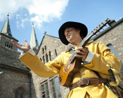 hx-jostziegenhirt-andreashub (4).jpg Ein mittelalterlich gekleideter Mann spielt Laute vor einer historischen Kirche an einem sonnigen Tag.