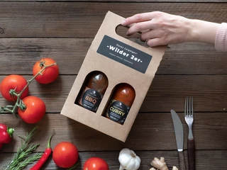 Kern & Stein Saucenbox "Wilder 2er" - Regionale Produkte aus Leipzig Eine Hand hält eine Pappverpackung mit 2 Feinkostsaucen der Kern und Stein Manufaktur aus Gohlis ins Bild, auf dem Tisch darunter liegen Tomaten, Paprika, Knoblauch und Chili, Regionale Produkte aus LeipzigA hand holding out a cardboard box with 2 bottles of sauce made by Kern & Stein in Gohlis. On the table below, you can see tomatoes, red peppers, garlic and chili, regional products from Leipzig