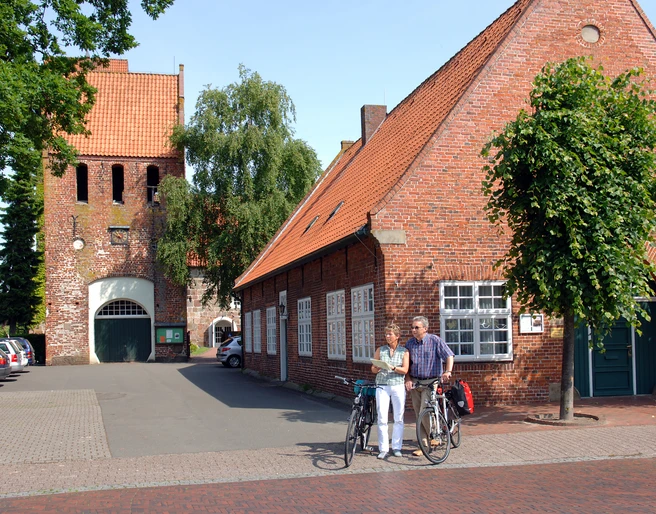 07_056.JPG Ein altes Backsteingebäude mit roten Dachziegeln und Bäumen davor. Zwei Radfahrer stehen lachend daneben.
