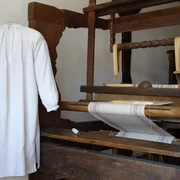 Das Hemd ohne Naht Ein historischer Webstuhl mit einem weißen, nahtlosen Hemd daneben, zeigt traditionelle Webkunst.