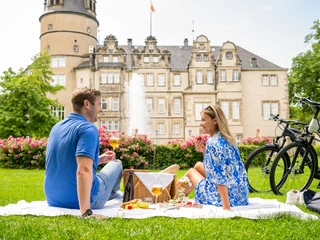 Detmold-2023-160-Residenzschloss-Schlosspark-Teutoburger-Wald-Tourismus-D-Ketz.jpg Ein Paar genießt ein Picknick auf einer Wiese vor einem historischen Schloss in Detmold.
