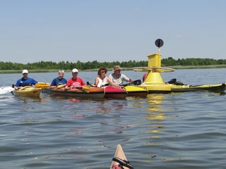 Fünf Kajakfahrer auf einem ruhigen See paddeln gemeinsam vor einer gelben Postboje unter blauem Himmel.