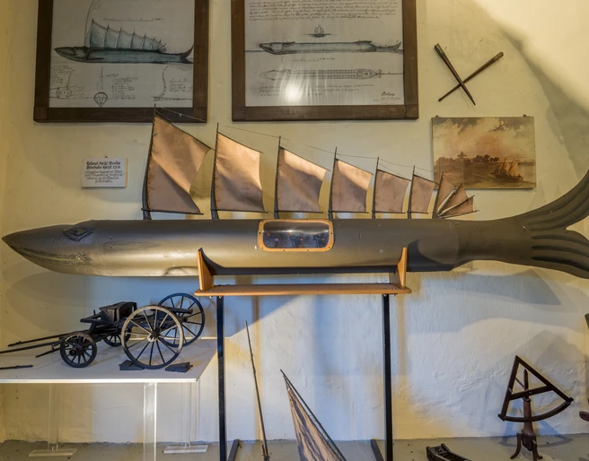 Modell eines Fisches mit Segeln, umgeben von Zeichnungen und nautischen Instrumenten an der Wand.