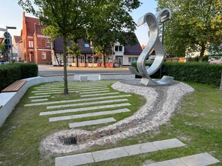 Wave Moderne Skulptur in urbaner Umgebung mit gepflastertem Weg und parkähnlicher Grünanlage.