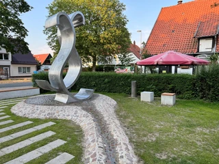 Metallene Skulptur einer geschwungenen Welle in einem kleinen Park, flankiert von Bäumen und Häusern.