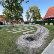 Wave Moderne Metallskulptur in einem grünen Parkbereich mit Bäumen und umliegenden historischen Gebäuden.
