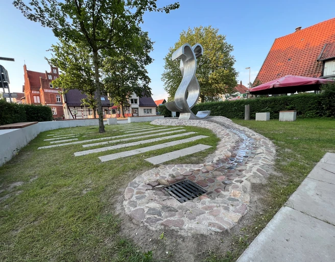 Wave Moderne Metallskulptur in einem grünen Parkbereich mit Bäumen und umliegenden historischen Gebäuden.