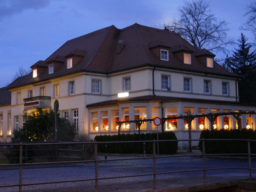 Zeitgeist Sächsisches Haus.JPG Großes, beleuchtetes Gebäude bei Dämmerung, mit roten Vorhängen und Weihnachtsdekoration in den Fenstern, umgeben von Bäumen.