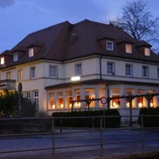Zeitgeist Sächsisches Haus.JPG Großes, beleuchtetes Gebäude bei Dämmerung, mit roten Vorhängen und Weihnachtsdekoration in den Fenstern, umgeben von Bäumen.