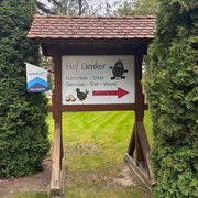 Hofladen Denker Hölzernes Schild des Hofladens Denker mit Hinweis auf Kartoffeln, Gemüse und Eier, umrahmt von Nadelbäumen.