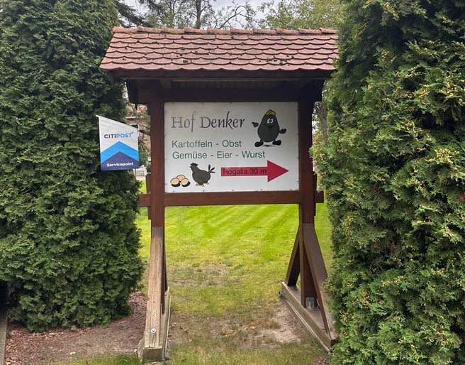 Hofladen Denker Hölzernes Schild des Hofladens Denker mit Hinweis auf Kartoffeln, Gemüse und Eier, umrahmt von Nadelbäumen.