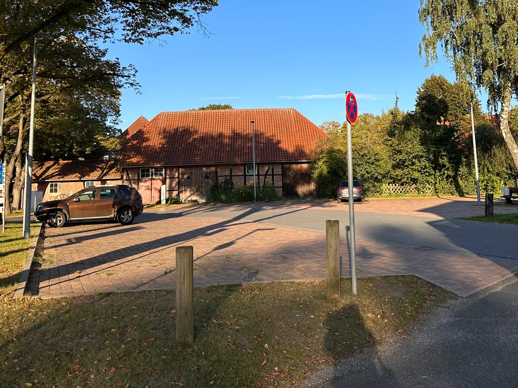 Parkplatz mit Pflastersteinen, umrahmt von Bäumen und einem Gebäude mit rotem Ziegeldach.