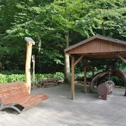 Hörstation Anna & Hermann in Bad Iburg Interaktive Hörstation mit Waldgeräuschen unter Holzpavillon nahe einer Ruhebank in einem bewaldeten Park.