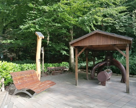 Hörstation Anna & Hermann in Bad Iburg Interaktive Hörstation mit Waldgeräuschen unter Holzpavillon nahe einer Ruhebank in einem bewaldeten Park.