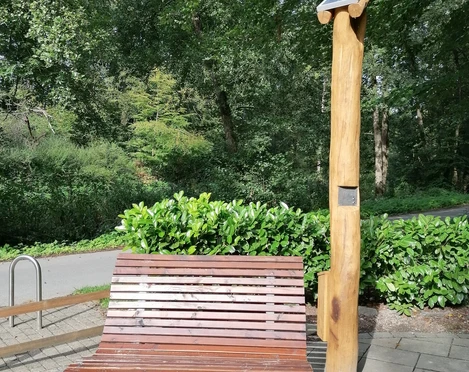 Hörstation Anna & Hermann in Bad Iburg Holzbank mit Rückenlehne neben einer naturholzfarbenen Hörstation in einem Waldbereich.