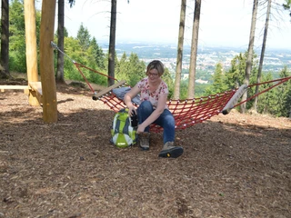 Hörstation Anna & Hermann in Werther Eine Frau sitzt in einer roten Hängematte im Wald, backpacker-rastend mit Ausblick auf eine Stadt.