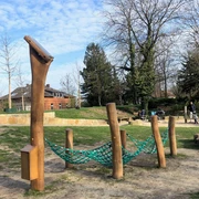Hörstation Anna & Hermann in Bielefeld Ein Spielplatz mit Kletternetz und Holzgestellen, umgeben von Bäumen und einem gepflasterten Weg.
