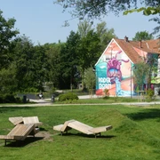 Graffiti von einem lila Monster auf einem Haus in Paderborn, umgeben von grünen Parkflächen.