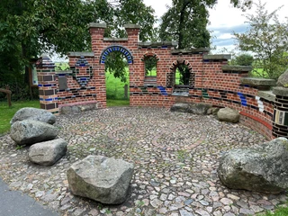 Altenbücken Backsteinskulptur mit bunten Fliesen in runder Anordnung, umgeben von Bäumen und großen Steinen.