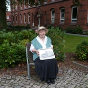 "Bürgerin Gesine" vor dem Gymnasium am Markt (früher Marktschule) Eine ältere Frau sitzt auf einer Bank, hält eine alte Fotografie vor einem historischen Schulgebäude.