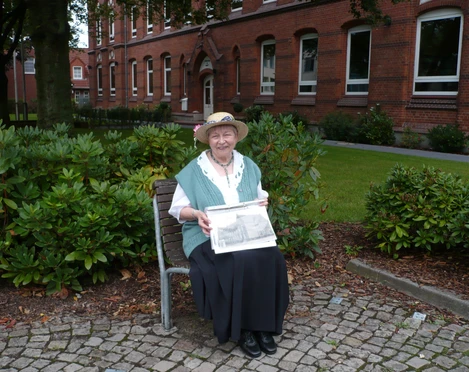 "Bürgerin Gesine" vor dem Gymnasium am Markt (früher Marktschule) Eine ältere Frau sitzt auf einer Bank, hält eine alte Fotografie vor einem historischen Schulgebäude.