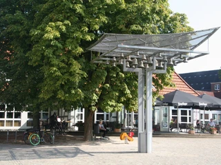 Ein modernes Glockenspiel steht auf einem gepflasterten Platz, umgeben von Bäumen und Menschen.